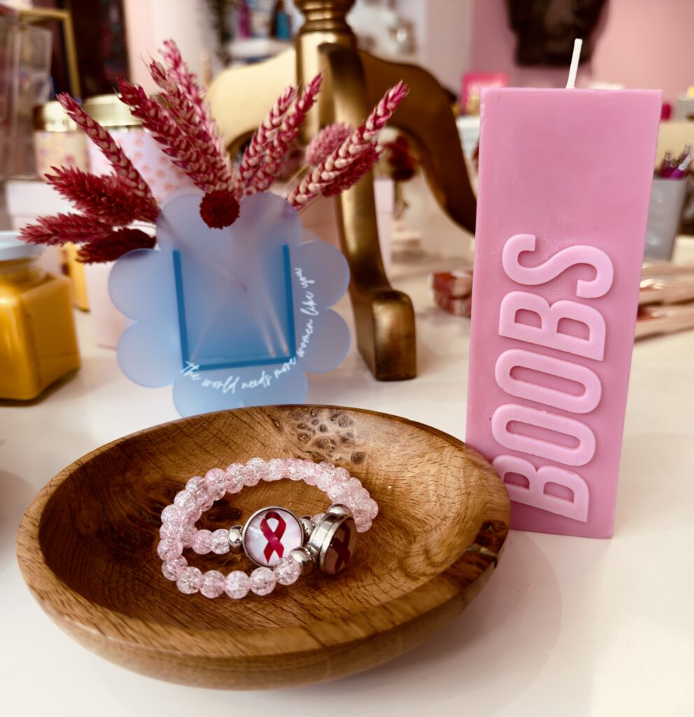 Still life with a dark wood bowl with a pale pink bracelet inside. Bracelet has the pink ribbon for breast cancer awareness. Next to the bowl is a pale pink candle with 'boobs' inscribed up the side.
