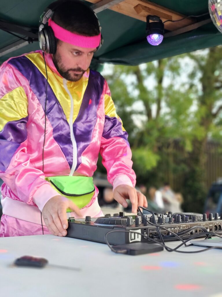 Man with a beard dressing a yellow, pink and purple shell suit style tracksuit with a digital mixing desk.