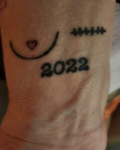 Image of a wrist tattoo, showing one breast and 2022, the breast as a scar line
